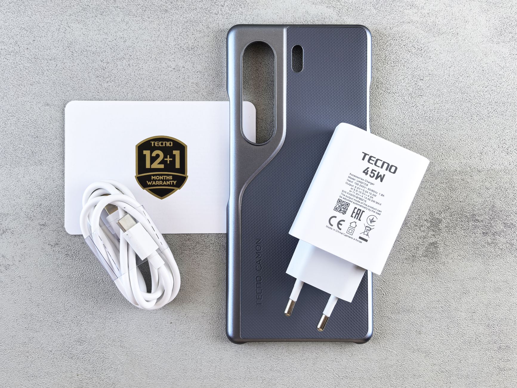 Flat lay of Tecno Camon phone case, charger, cable, and warranty card on a textured surface.