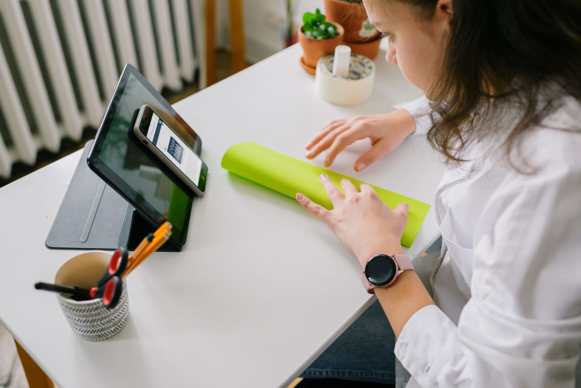 A woman using tablet and smartphone at a desk, arranging a roll of paper.