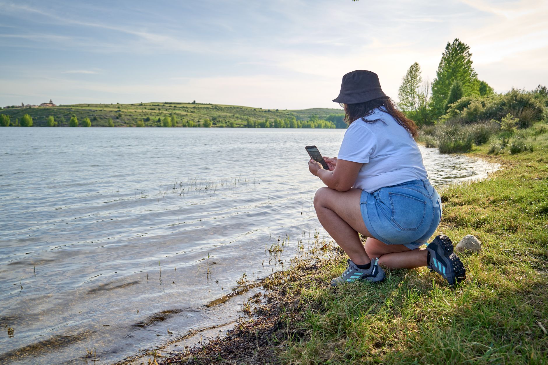 Woman kneeling by the riverbank in Segovia, capturing scenic nature photos with her phone.