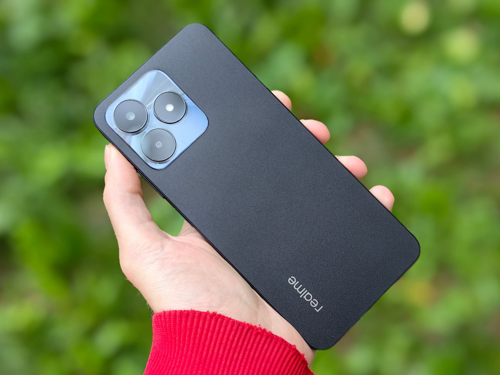 A close-up shot of a hand holding a Realme smartphone with a blurred green background, showcasing modern technology outdoors.