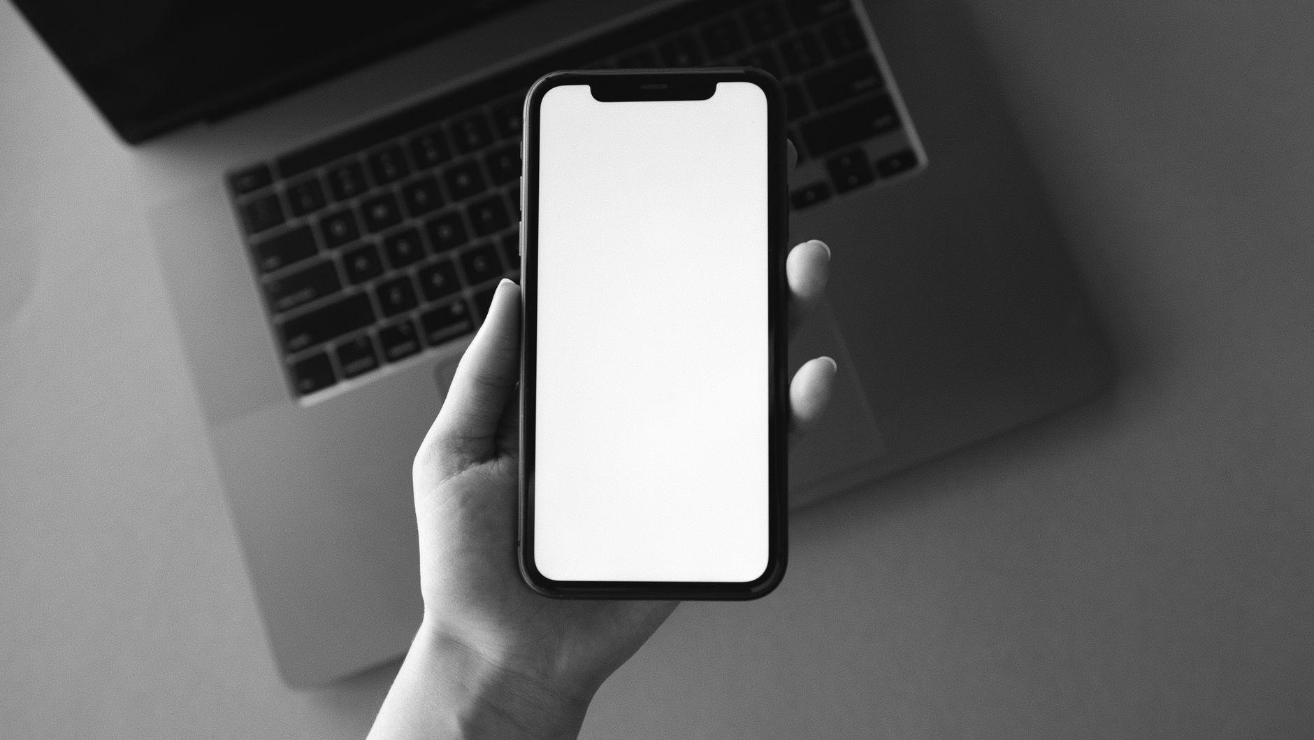 Monochrome image of a hand holding a blank smartphone screen above a laptop, ideal for tech presentations.