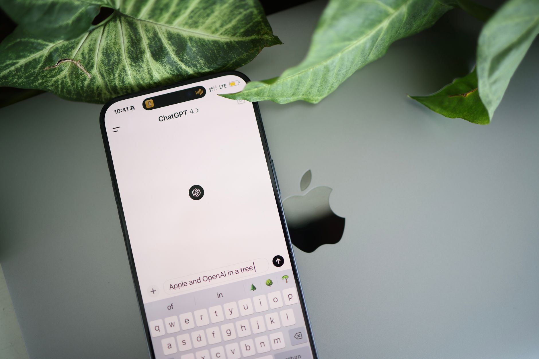 A smartphone shows a ChatGPT interface placed on an Apple laptop in a leafy environment.