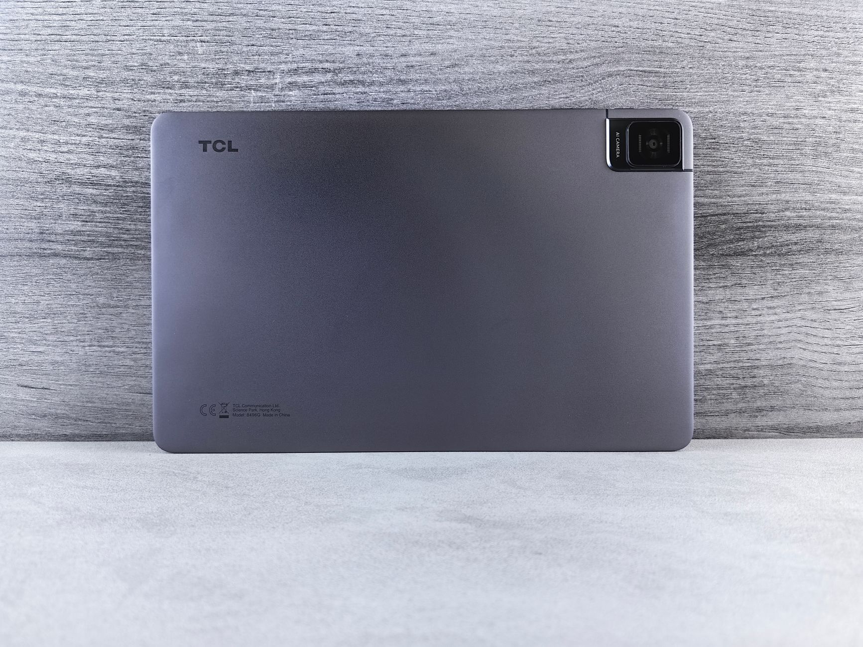 Gray TCL tablet displayed on a minimalist surface with a neutral gray wooden background.